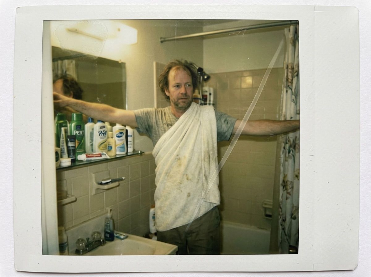A man poses like a statue in a small bathroom with a towel over one shoulder