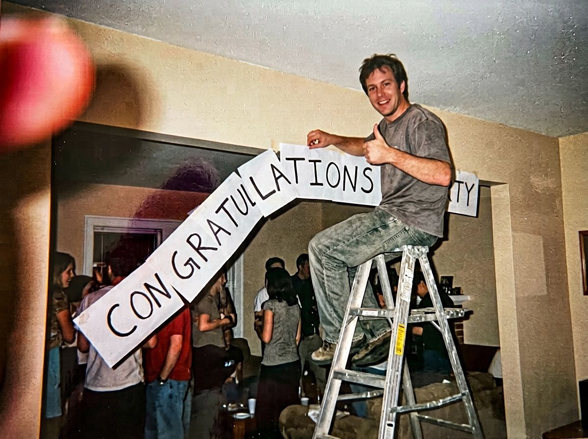 A man on a stepladder gives a thumbs up under a half-fallen banner reading CONGRATUL