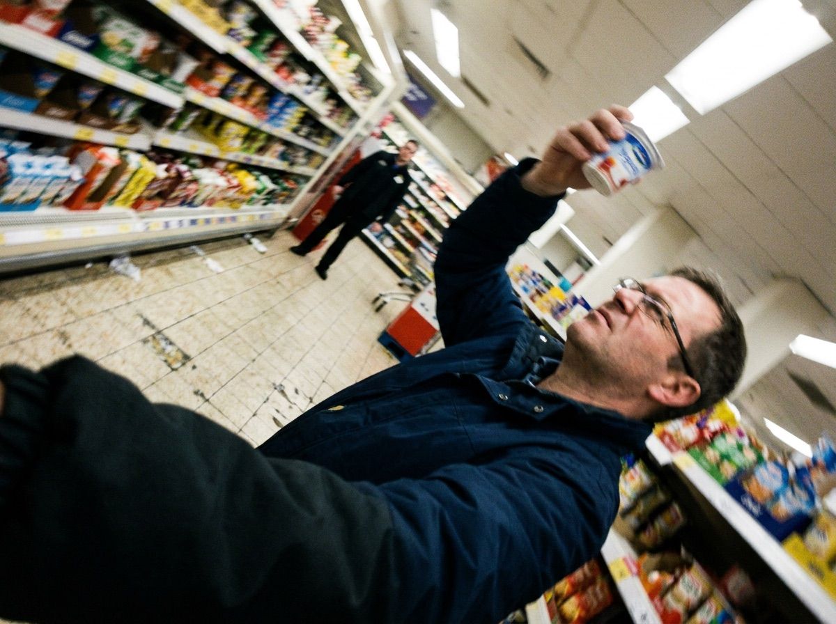 A man inspects a yoghurt date while a shop employee watches from the end of the aisle