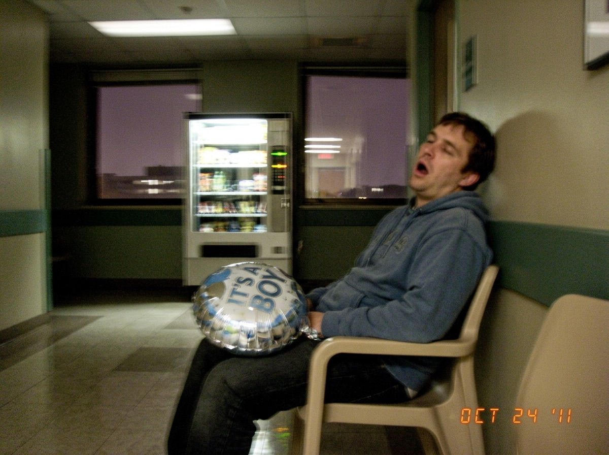 A man asleep in a hospital corridor chair with a deflating IT'S A BOY balloon on his lap