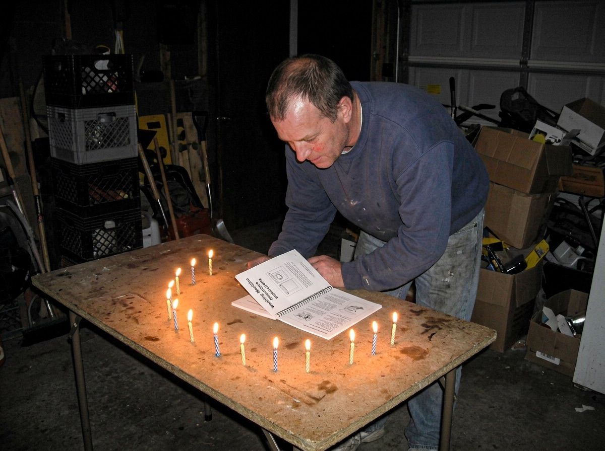 A man reads a washing machine manual by the light of nine candles arranged on a garage table