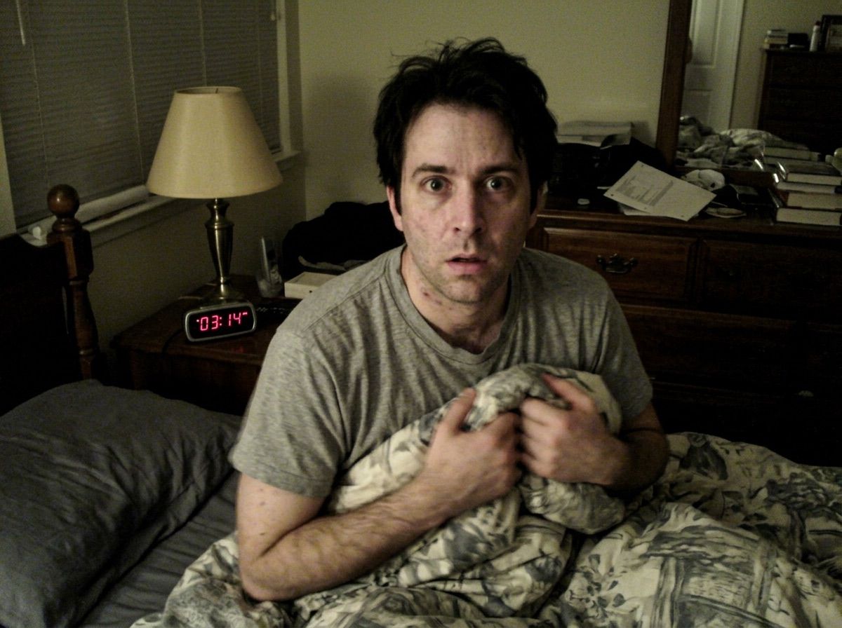 A man sits bolt upright in a fully lit bedroom at 3:14 in the morning