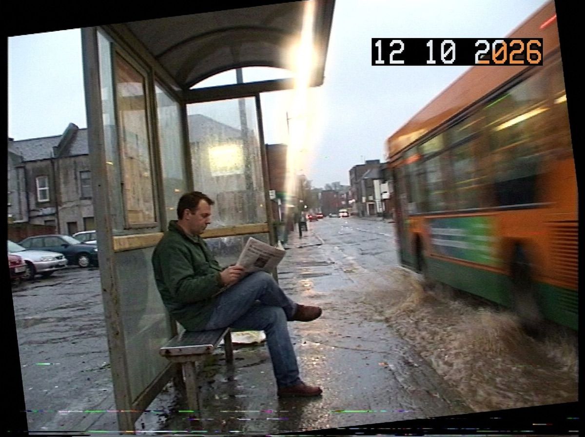A man reads a newspaper at a bus stop as a wall of puddle water hangs in the air beside him