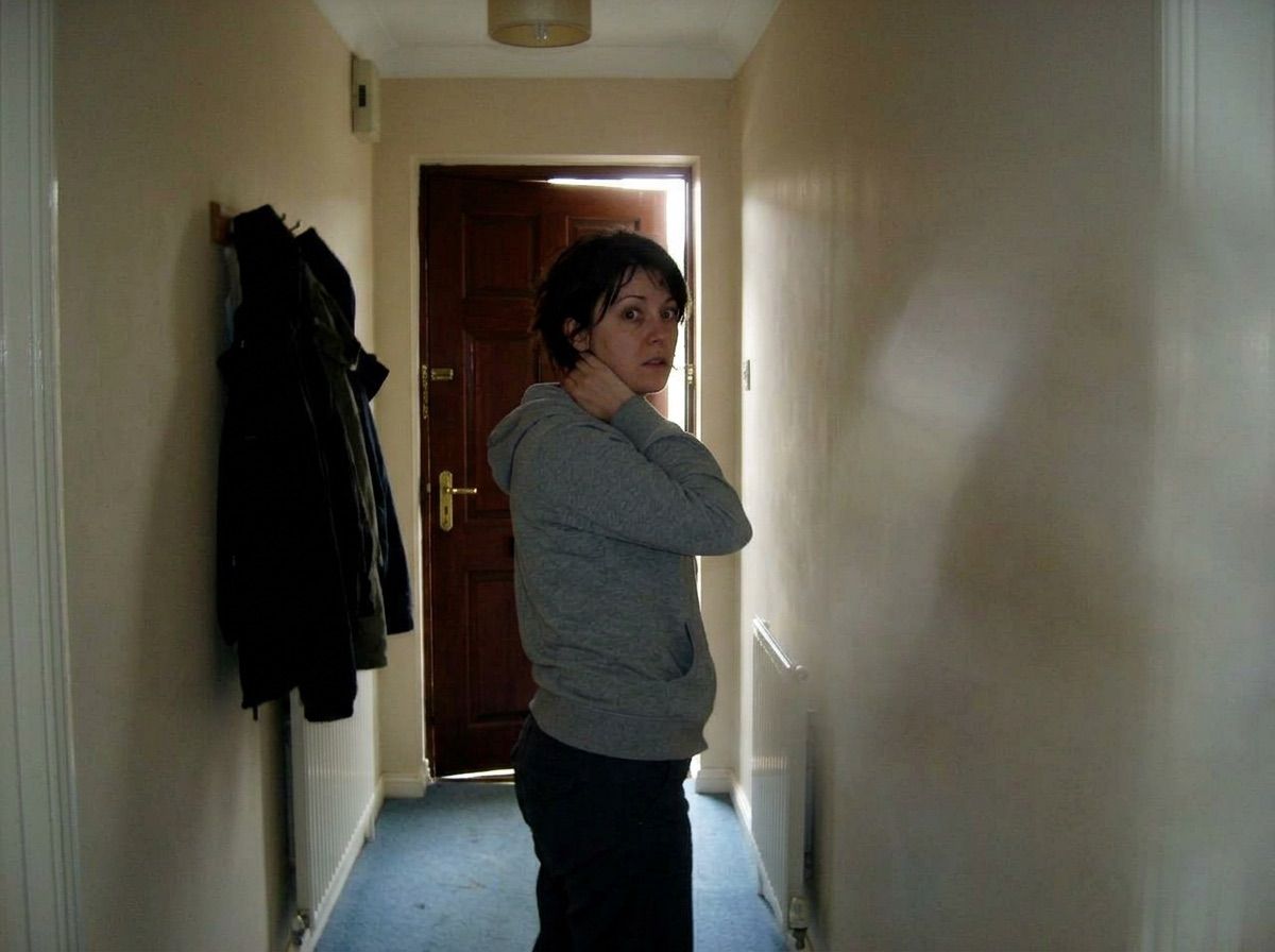 A woman touches the back of her neck in a hallway with the front door slightly ajar