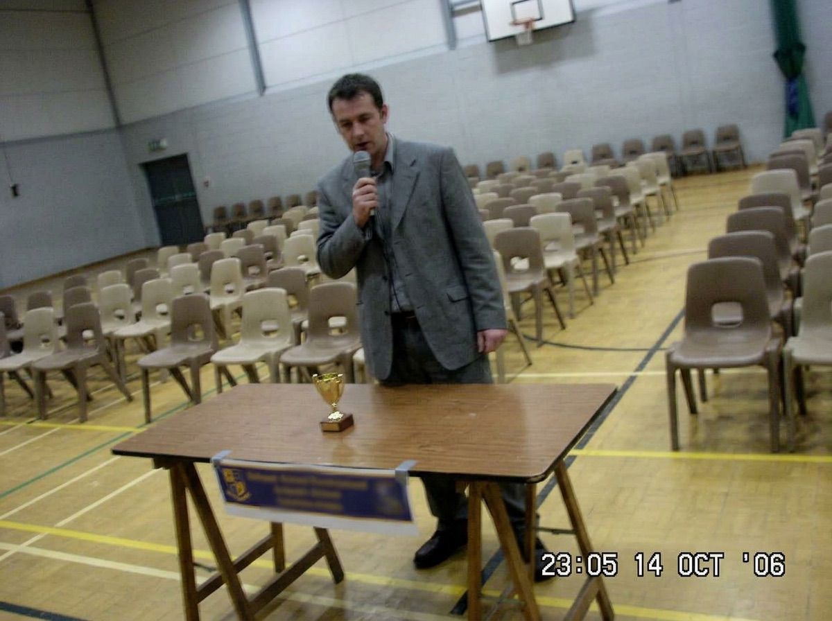 A man with a microphone beside a trophy table holding one small trophy in an empty hall