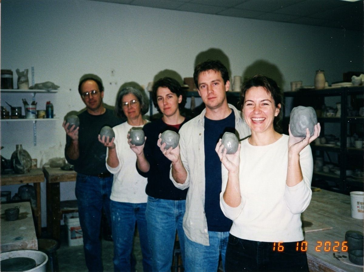 Six people at a pottery class hold up near-identical grey lumps of clay